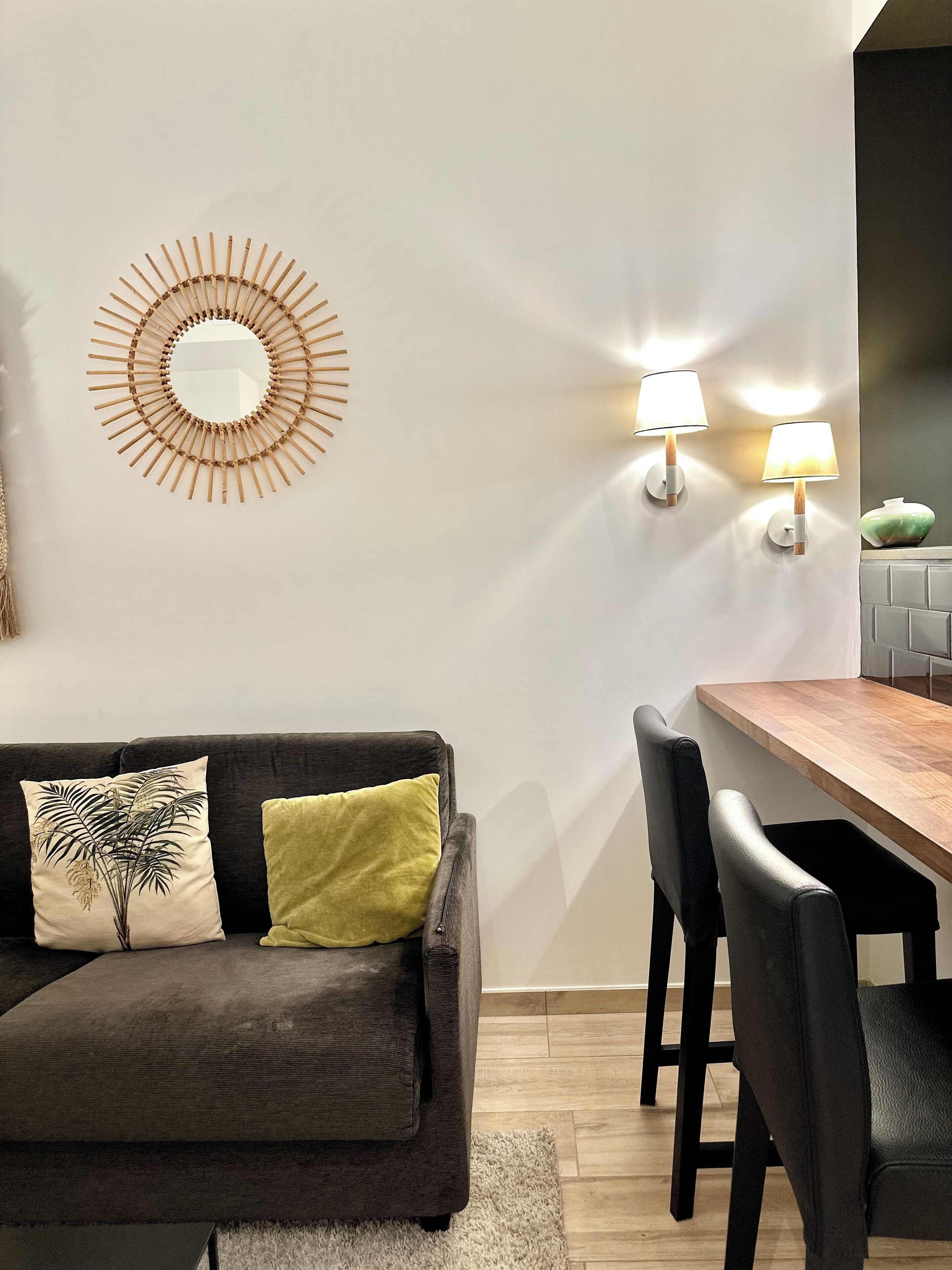 Una sala moderna con un sofá gris oscuro, dos cojines, un espejo redondo en la pared y una mesa de comedor de madera con sillas.