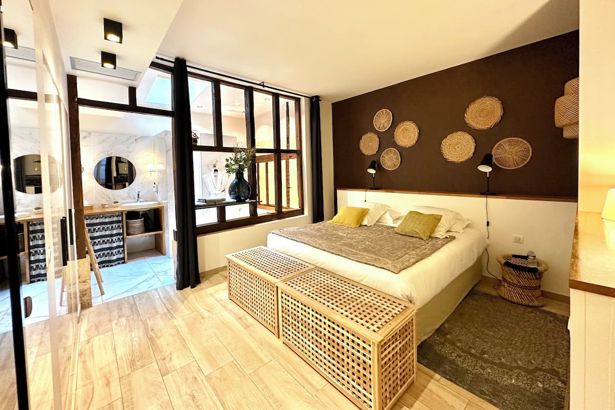 A modern bedroom with a large bed, a wooden bench, a view of the bathroom, and decorative woven wall art.