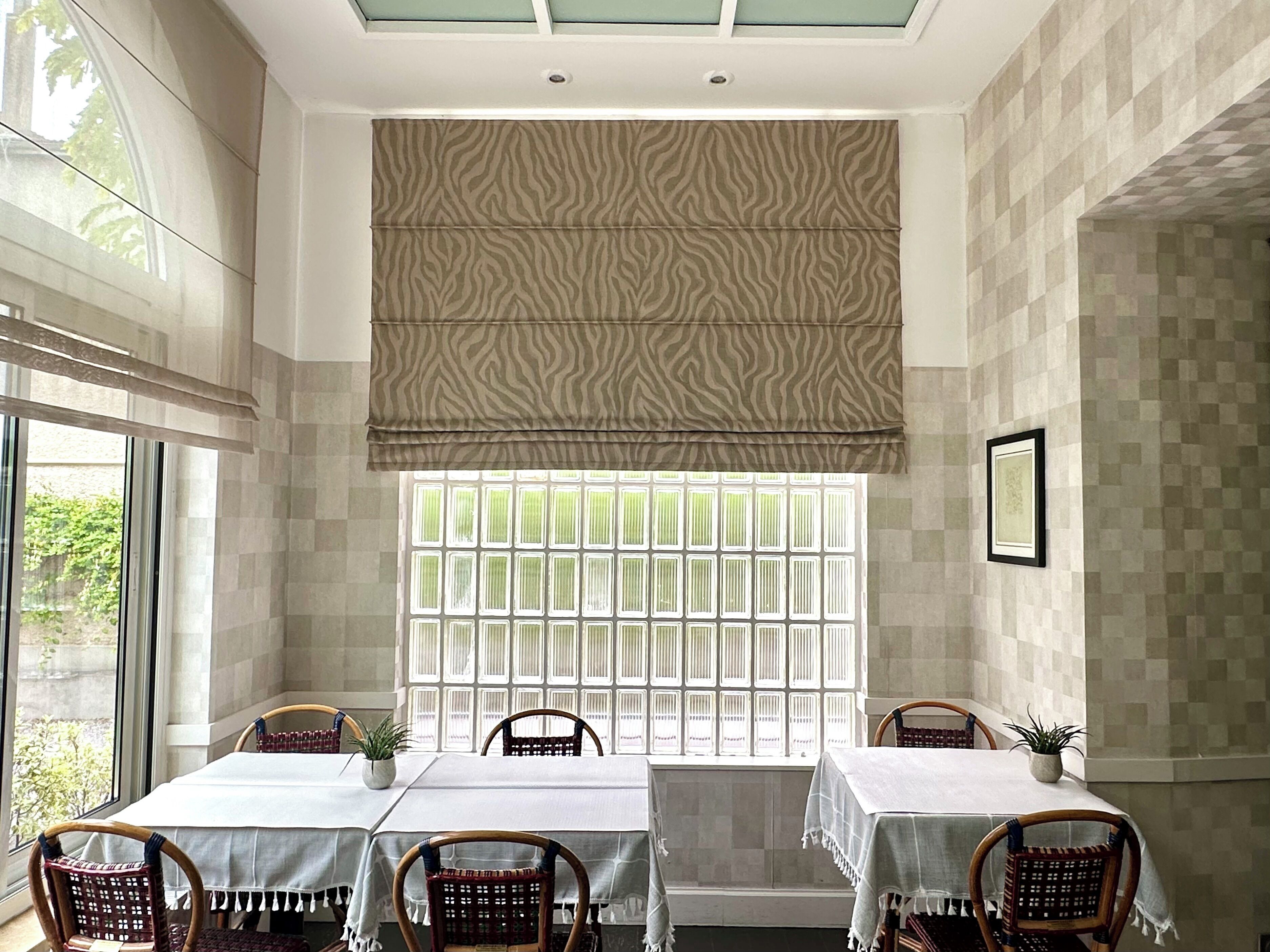 A dining area with a table set for four, surrounded by wicker chairs, a window with a patterned blind, and a framed picture on the wall.