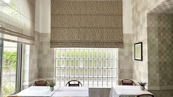 A dining area with a table set for four, surrounded by wicker chairs, a window with a patterned blind, and a framed picture on the wall.