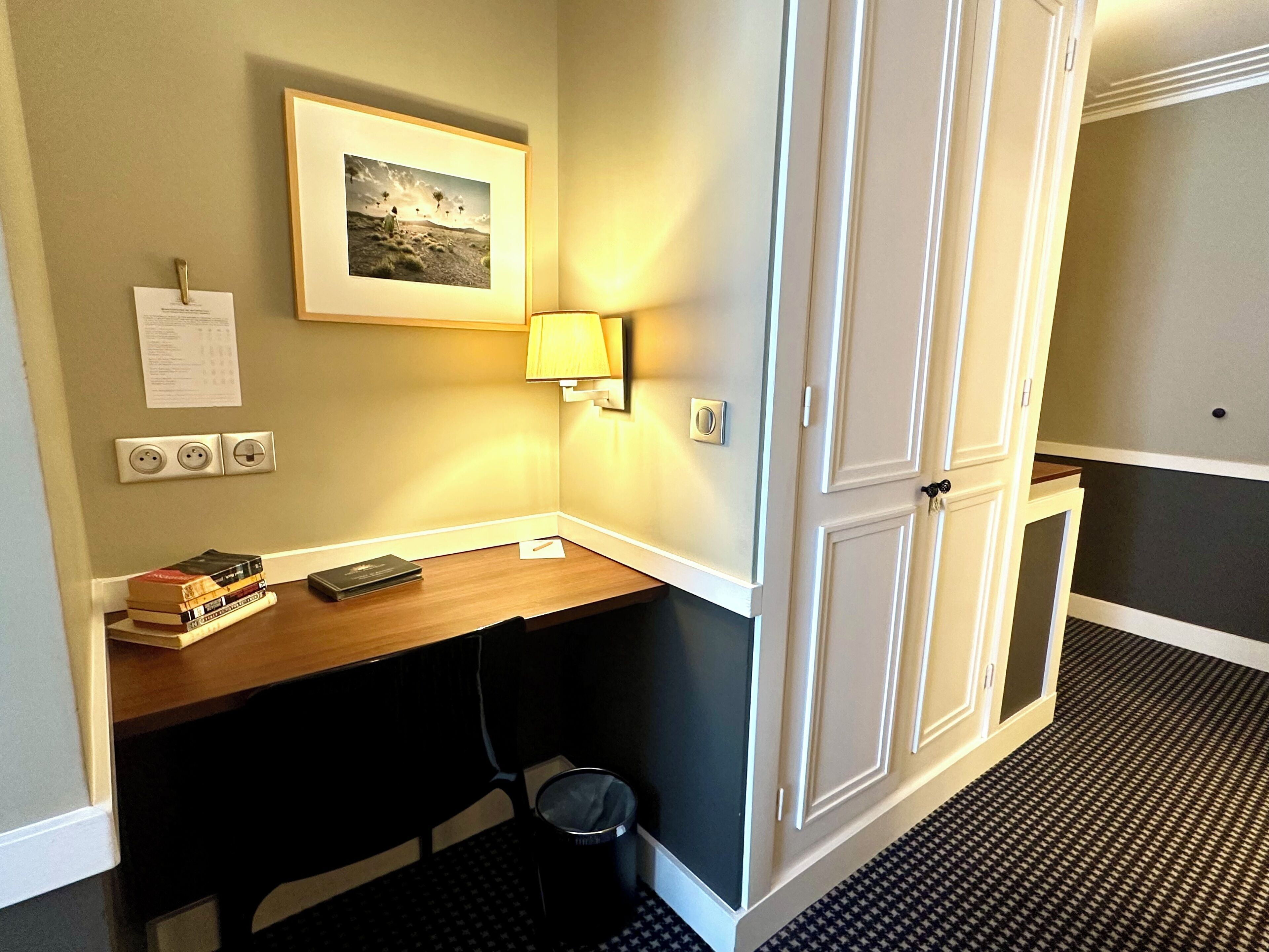 A hotel room with a desk, a lamp, a picture on the wall, a trash bin, and a door.