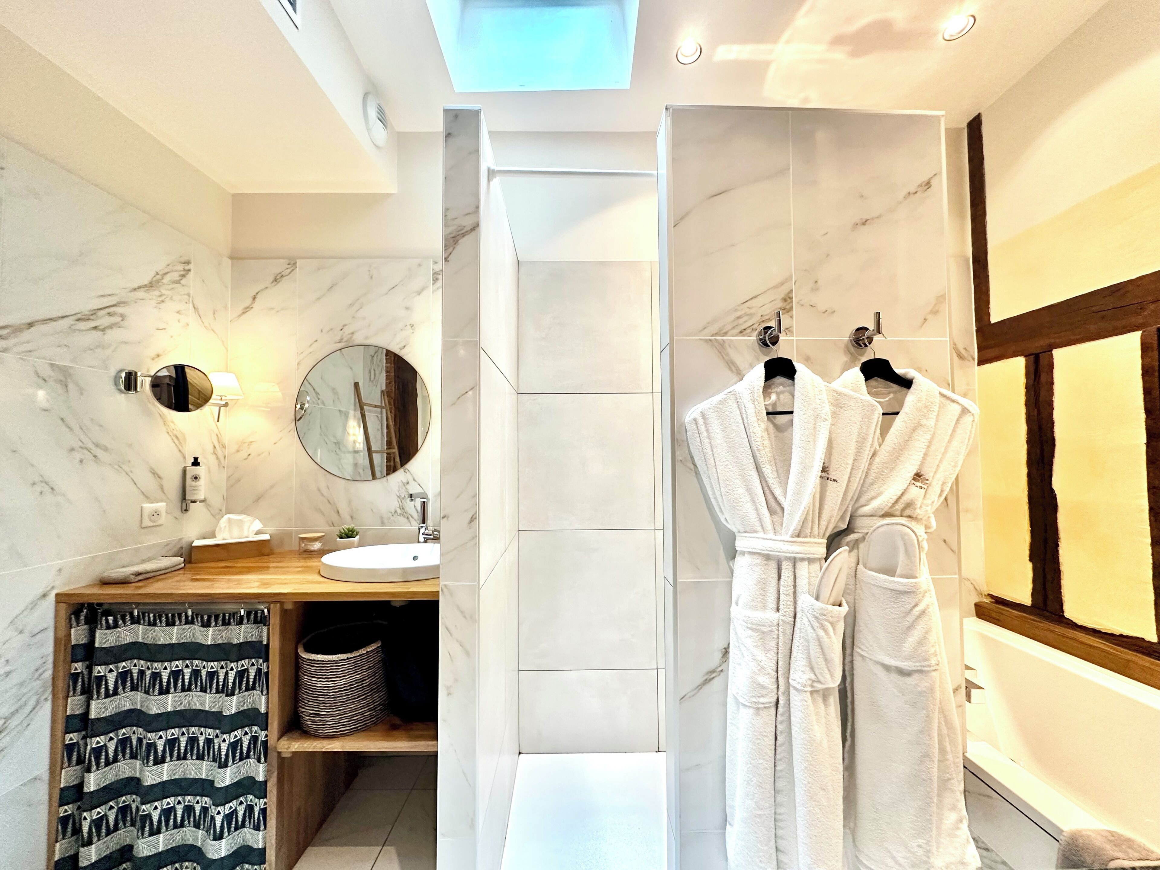 Un baño moderno con tocador de mármol, espejo redondo, toallero con dos albornoces blancos y un tragaluz sobre la zona de la ducha.