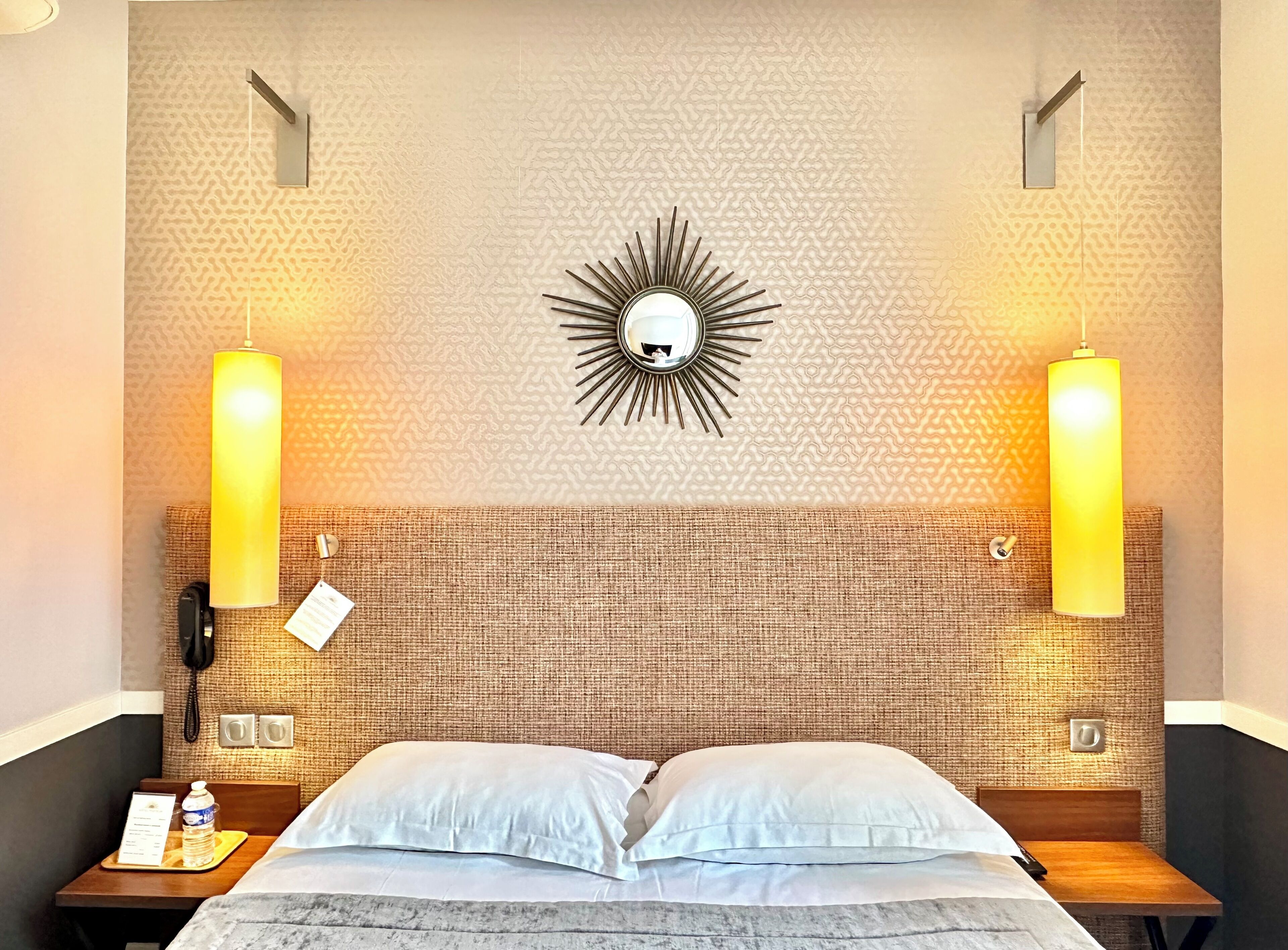 Une chambre d’hôtel avec un mur à motifs, un miroir en forme de soleil, deux lampes fixées au mur, une table de chevet avec un téléphone, une bouteille d’eau et un plateau avec un menu.