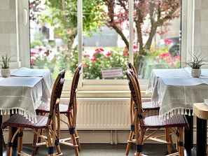 A dining area with wicker chairs and tables, a view of a garden with pink flowers through a window. - MAISON LIA  (Châlons-en-Champagne)