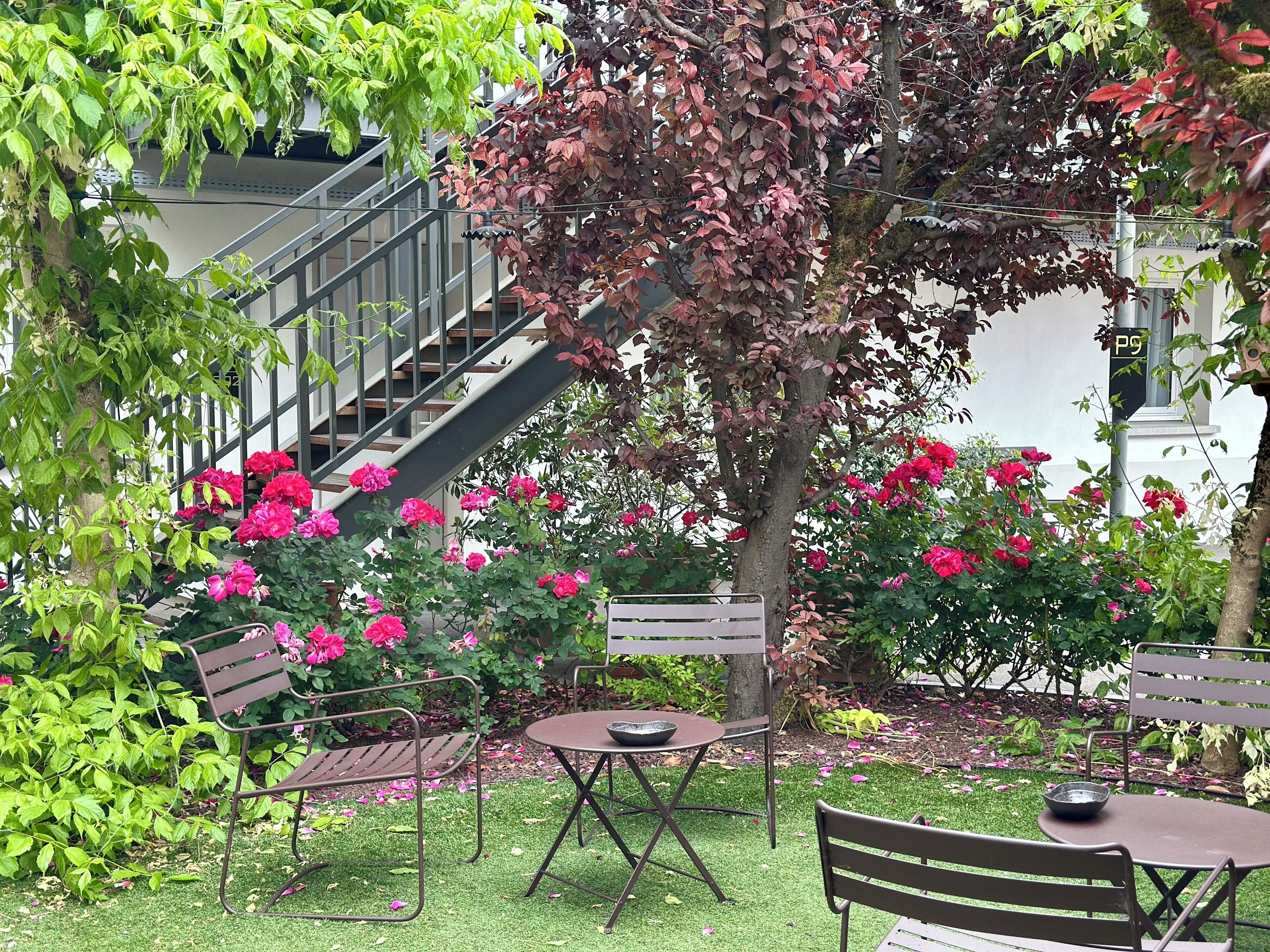 Un espacio de estar al aire libre con sillas de metal y una mesa redonda, rodeado de exuberante follaje y plantas con flores.