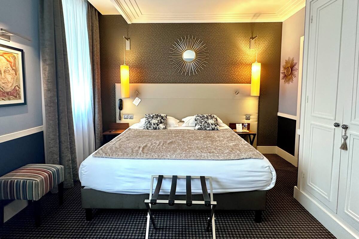 A hotel room with a large bed, bedside lamps, a wall clock, and a small table with a phone and a bottle.
