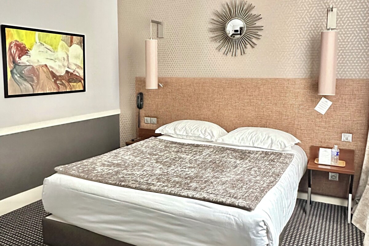 Une chambre d’hôtel avec un grand lit, une tête de lit, un miroir fixé au mur, un miroir décoratif en forme de soleil, deux lampes de chevet, un tableau encadré et une petite table avec une bouteille d’eau.