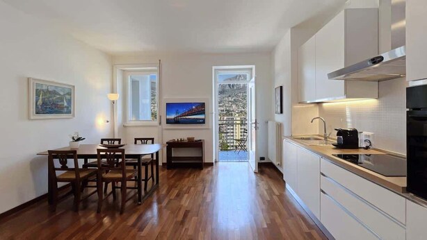 Eine moderne Küche mit Holzboden, einem Essbereich mit Tisch und Stühlen, einem Fernseher und einem Balkon mit Blick auf die Stadt.