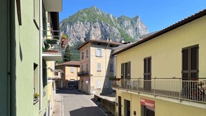 A narrow street with buildings on either side, a balcony with plants, and a mountain in the background. - Welcome House - Affitti Brevi italia (Lecco)
