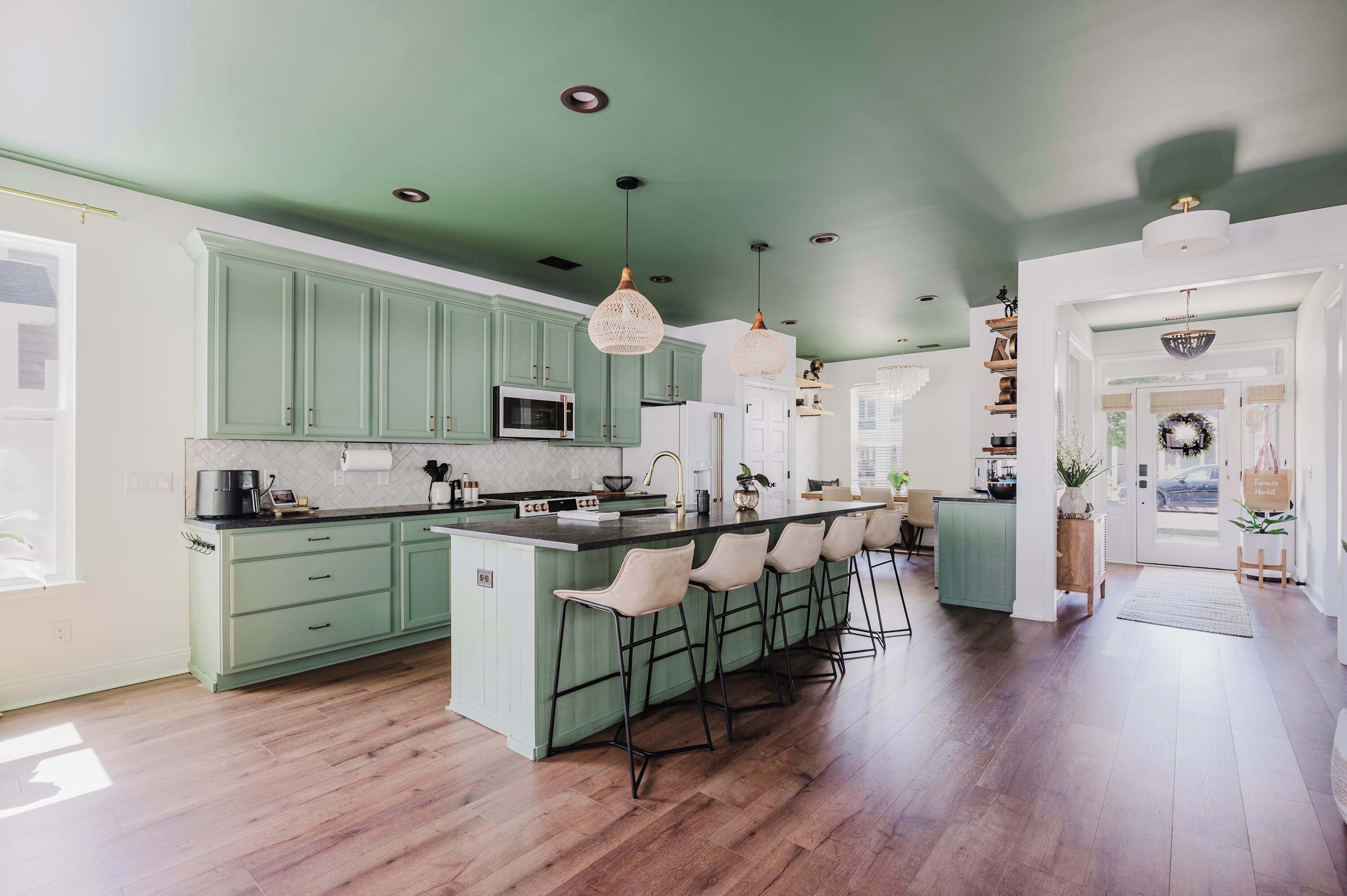 A kitchen with green cabinets, a central island with bar stools, pendant lighting, and a view of the dining area.