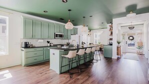 A kitchen with green cabinets, a central island with bar stools, pendant lighting, and a view of the dining area.