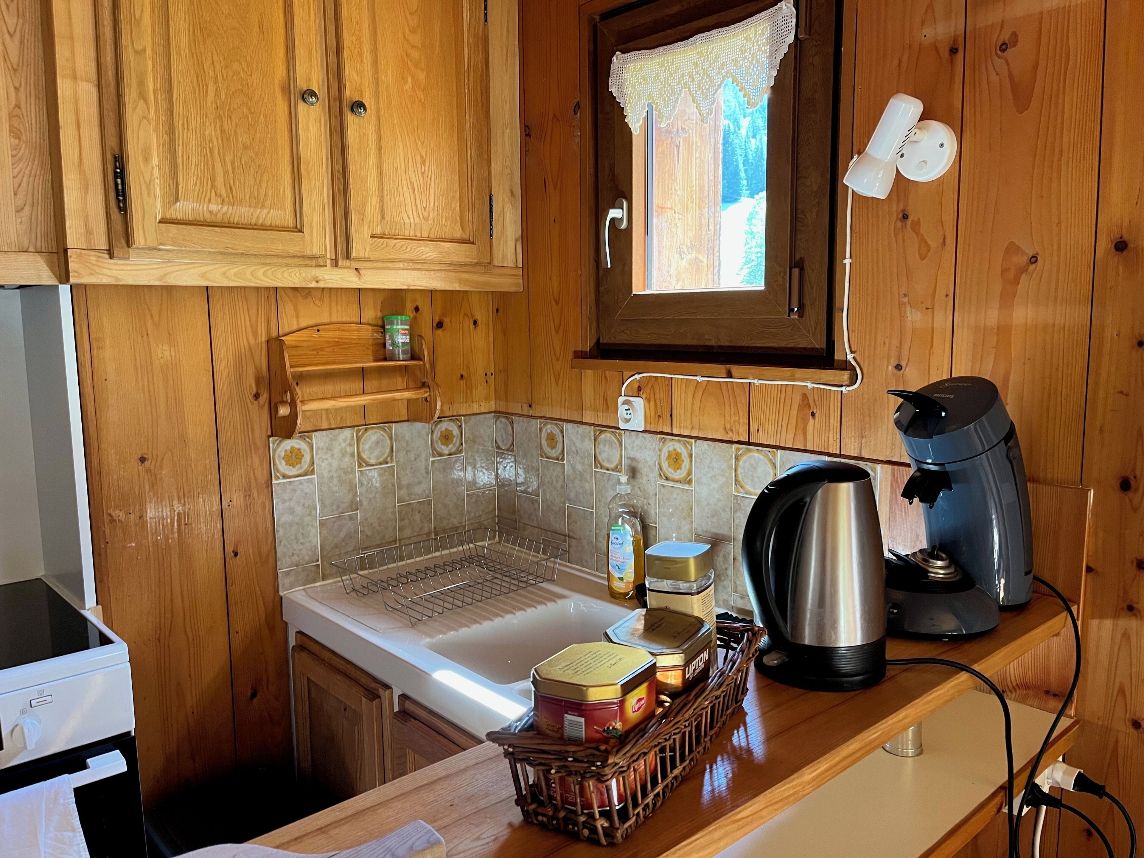 A wooden kitchen with a sink, a coffee maker, and a window with a lace curtain.