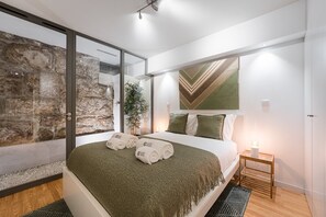 Une chambre moderne avec un grand lit, un mur en pierre et une vue sur l’extérieur, visible à travers une porte vitrée.