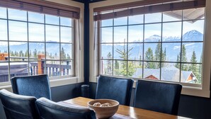 Un coin repas avec une table et des chaises, un saladier en bois, et une vue sur les montagnes à travers de grandes fenêtres.