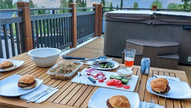 Une terrasse en bois avec une table dressée pour un repas, où l’on peut déguster des hamburgers, des salades et des boissons, le tout avec une vue imprenable sur un paysage montagneux.
