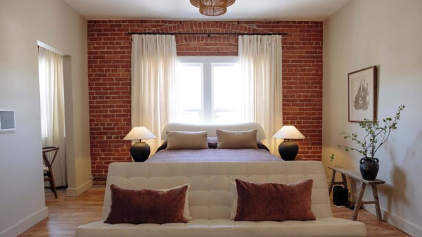 Ein Schlafzimmer mit Backsteinwand, einem Bett mit zwei Kissen, einem weißen Sofa und einem Fenster mit Vorhängen.