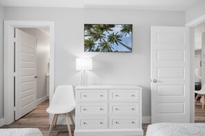 Una habitación con un aparador blanco, una silla, una lámpara y un cuadro con palmeras en la pared.