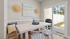Een eetruimte met een witte tafel, witte stoelen en een bank met kussens. Twee hoeden hangen aan de muur en er hangt een schilderij boven de tafel.