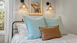 Een slaapkamer met een bed, gekleurde kussens, een kunstwerk in een lijst en wandlampen.