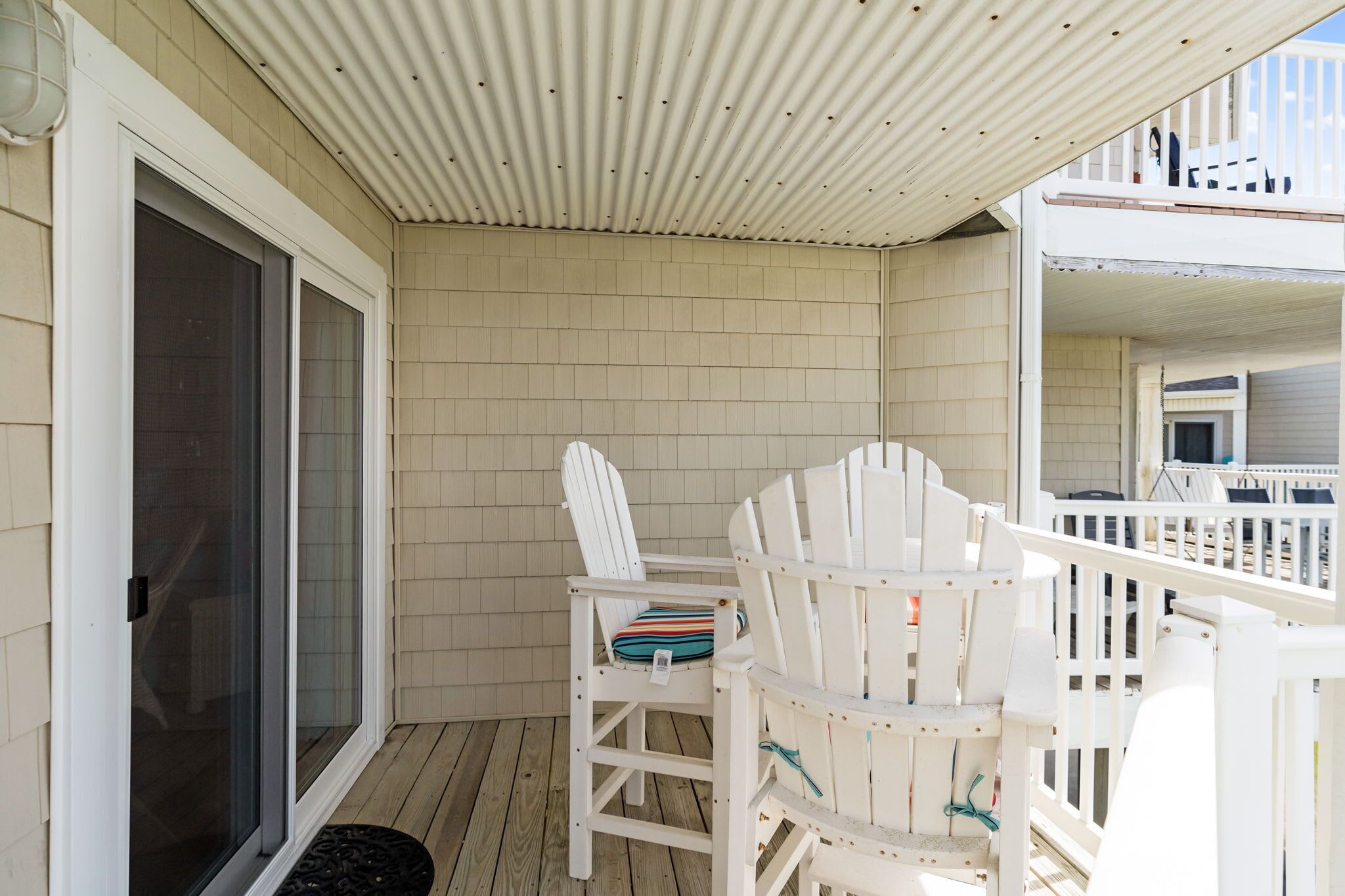 A covered outdoor area with a wooden deck, white Adirondack chairs, and a sliding glass door.