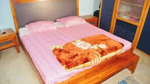 A single-bed room with a wooden headboard, a pink bedspread, and a floral-patterned blanket. There is a small wooden nightstand with a lamp, and a window with a curtain. - CHEZ TITI: SAFETY & COMFORT IN ST LOUIS (Saint-Louis)