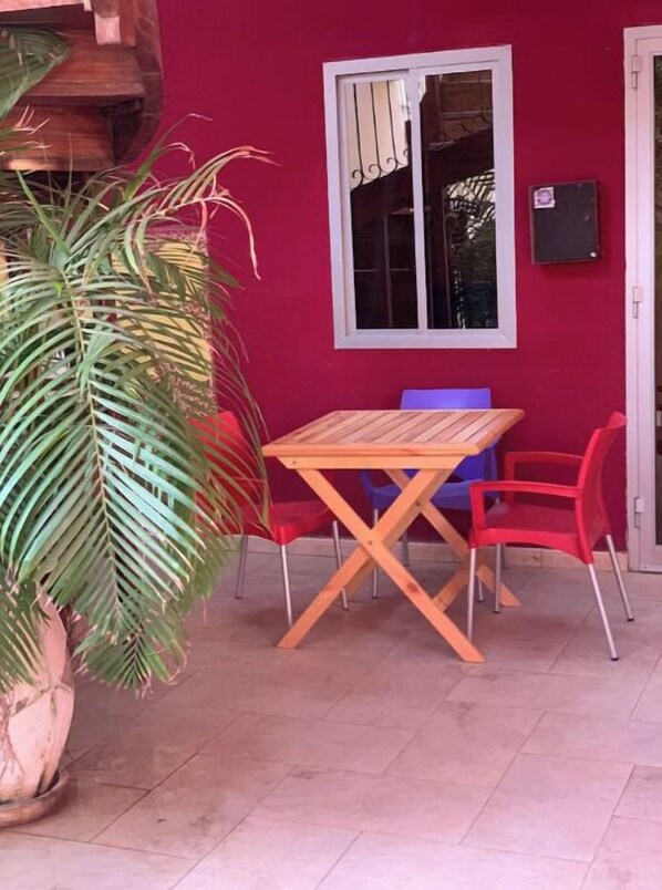 Une table et des chaises en bois sont placées devant un mur rouge. Une fenêtre se trouve au-dessus, et un pot de fleurs est posé sur le côté gauche.