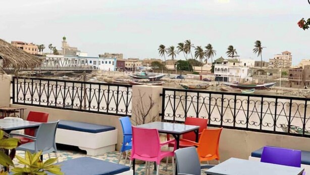 A rooftop terrace with colorful seating and tables, overlooking a cityscape with buildings and palm trees. - CHEZ TITI: SAFETY & COMFORT IN ST LOUIS (Saint-Louis)