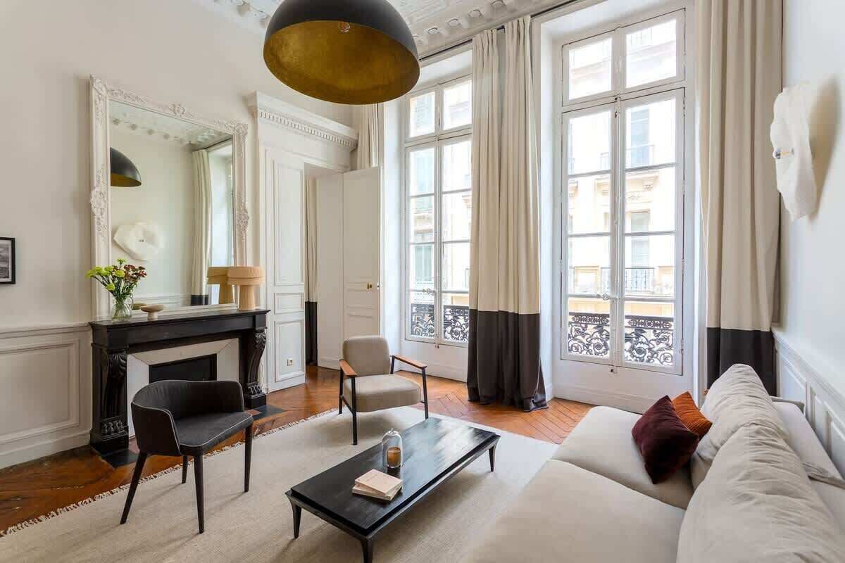 A living room with a fireplace, a sofa, a coffee table, and a chair. There is a large window with curtains and a view of buildings outside.
