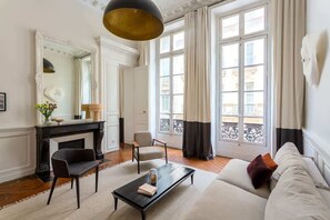 A living room with a fireplace, a sofa, a coffee table, and a chair. There is a large window with curtains and a view of buildings outside.