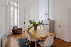 A dining area with a wooden table, white chairs, a potted plant, a brown leather chair, a floor lamp, and a refrigerator.