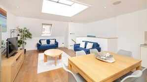 A modern living room with a wooden dining table, two blue sofas, a round coffee table, and a TV on a wooden stand.