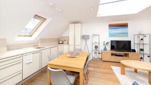 A modern kitchen with a skylight, a dining area with a wooden table, and a living area with a TV and shelving.