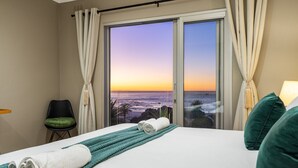 A hotel room with a bed, a view of the ocean at sunset, a green chair, and a teal throw blanket.