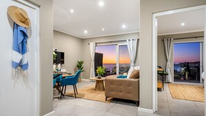 A modern living room with a sofa, dining area, and a view of the sunset through large windows.