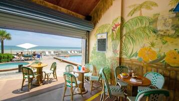 Een restaurant aan het strand met terras, een zwembad en een muurschildering van tropische bloemen en palmbomen.