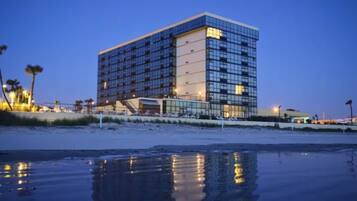 Een modern hotel met een glazen gevel, nabij een strand met een spiegelend wateroppervlak.
