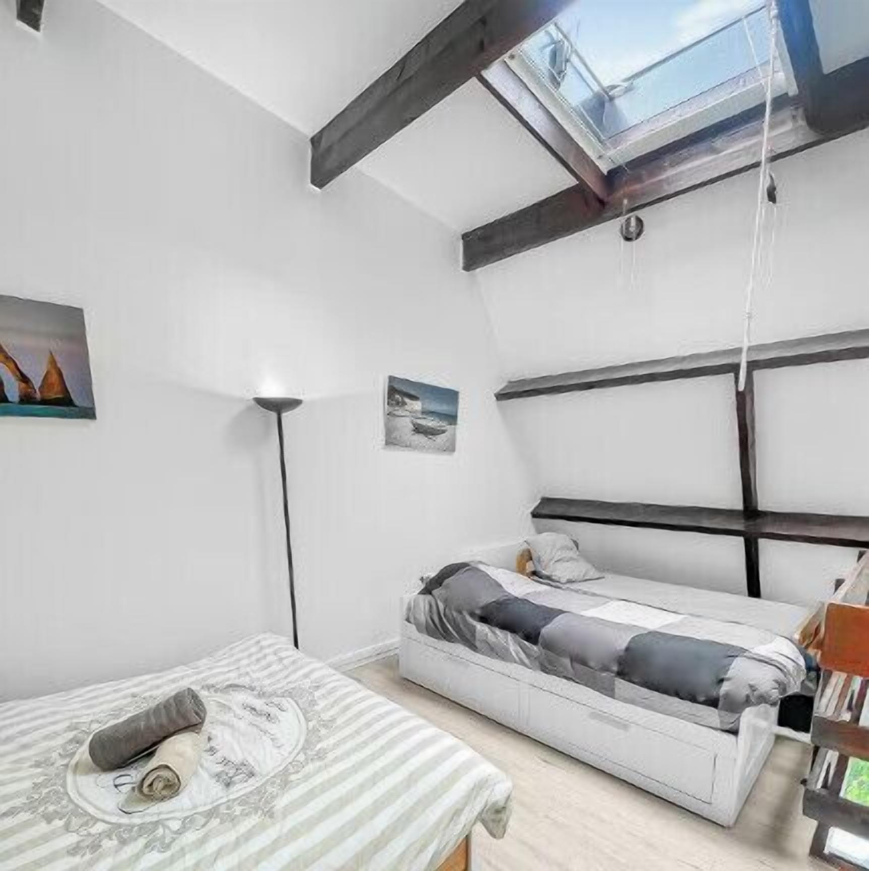 Une chambre à coucher de taille réduite, dotée d’une lucarne, d’un lit simple, d’une table de chevet et d’une lampe sur pied.