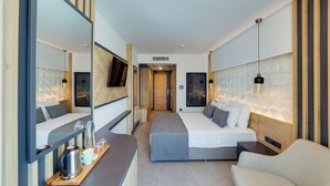 A modern hotel room with a large bed, a sofa, a desk with a kettle, and a mirror. - Signature Blue Resort (Kuşadası)
