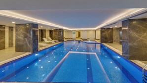 A large indoor swimming pool with a tiled floor and walls, surrounded by marble pillars and lounge chairs. - Signature Blue Resort (Kuşadası)