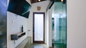 A hallway with a wooden bench, a mounted television, a wine bottle, and a door leading to a glass-walled room.