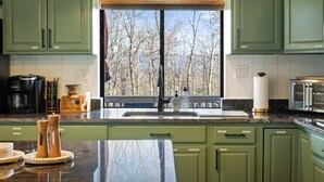 Une cuisine avec des meubles de couleur verte, un plan de travail en granit, une machine à café, un mixeur et une vue sur les arbres par la fenêtre.