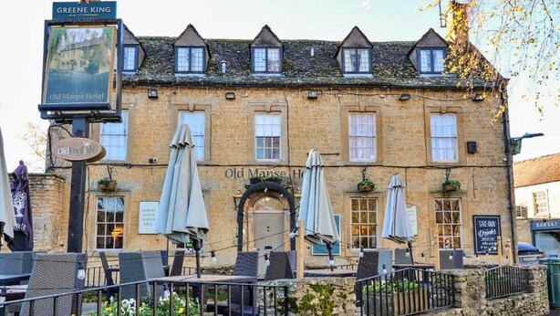 A traditional stone building with a sign for 'Old Manse Hotel' and 'Greene King' above it. There are outdoor tables and umbrellas, and a stone wall in front. - Holiday Home in Cotswolds With Hot Tub (Cheltenham)