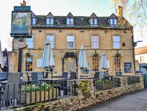 A traditional stone building with a sign for 'Old Manse Hotel' and 'Greene King' above it. There are outdoor tables and umbrellas, and a stone wall in front. - Holiday Home in Cotswolds With Hot Tub (Cheltenham)
