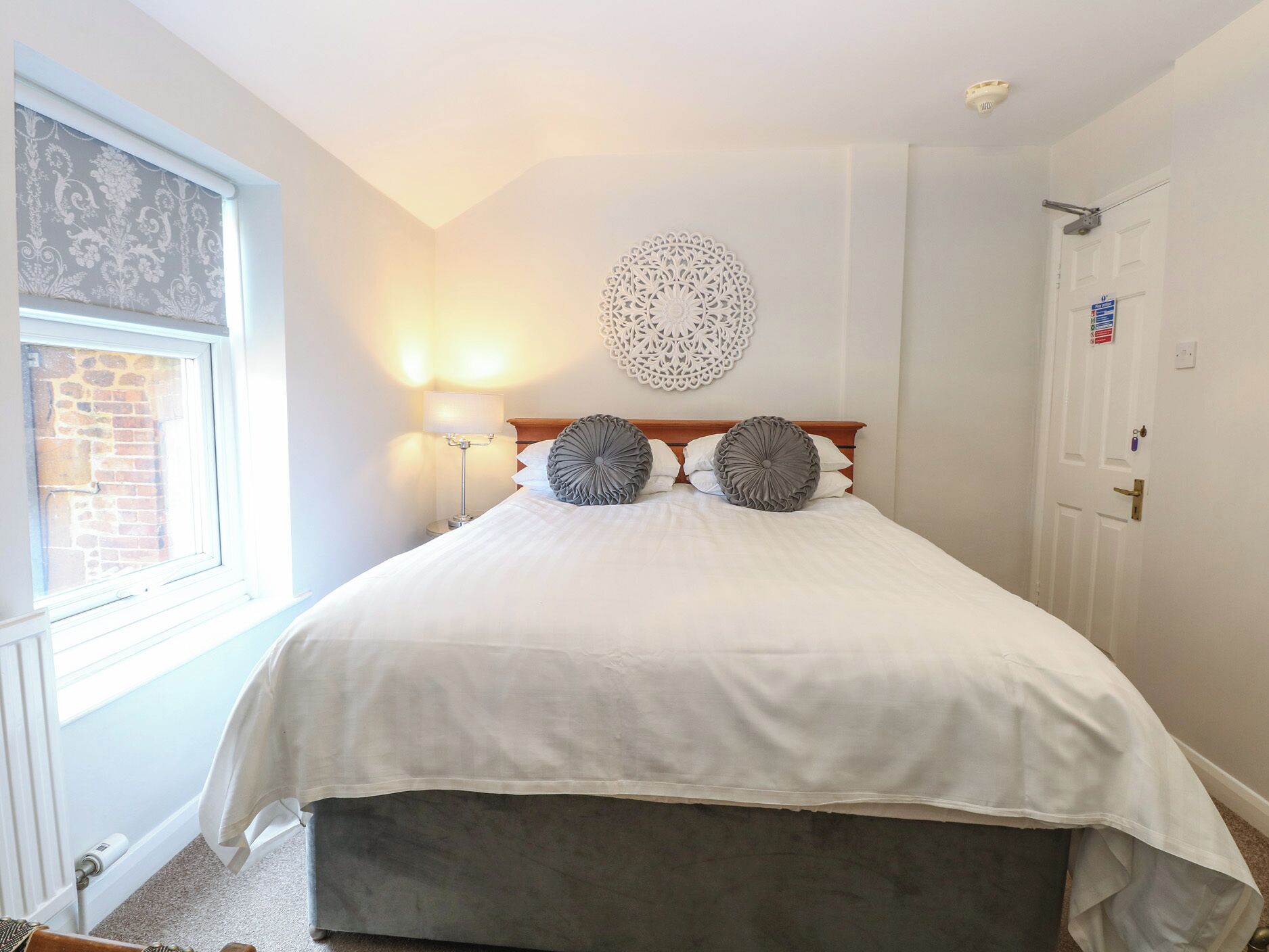 Un lit bien fait, avec une couette blanche et deux coussins décoratifs, un luminaire fixé au mur et une porte avec une pancarte.
