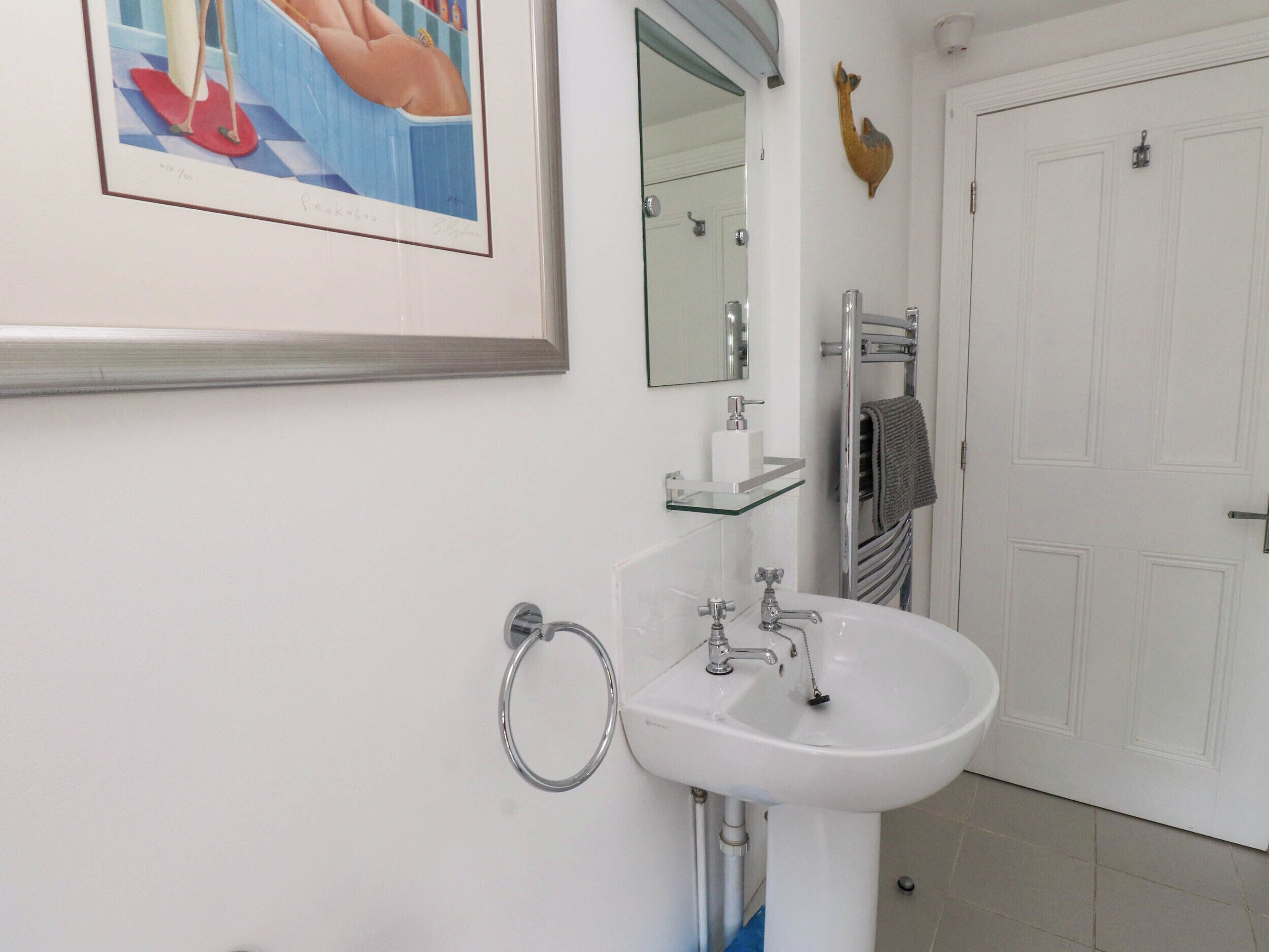 Une salle de bain avec un lavabo blanc, une étagère en verre, un porte-serviettes et un tableau encadré au mur.