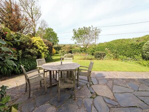 A stone patio with a round wooden table and chairs, surrounded by lush greenery and a well-maintained lawn. - Bryn Tirion (Pwllheli)