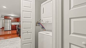 A laundry room with a white washer and dryer, a control panel, and a door leading to a kitchen area.
