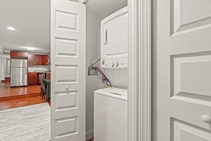 A laundry room with a white washer and dryer, a control panel, and a door leading to a kitchen area.