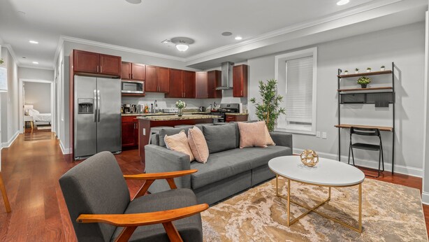 Een moderne woonkamer met een grijze bank, houten stoelen, een salontafel en een keuken op de achtergrond.