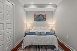 A hotel room with a bed, two bedside lamps, a painting of a canal, and a door.